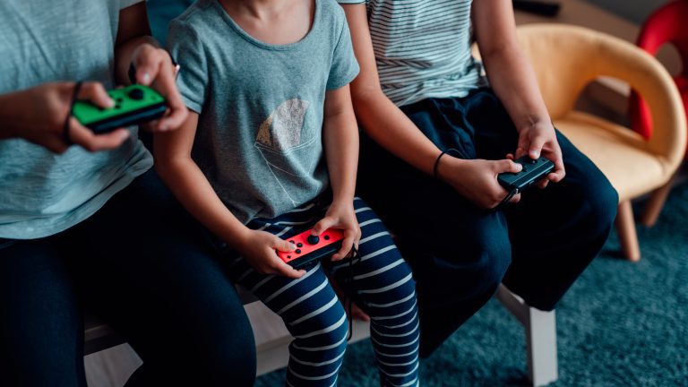 Drei jüngere Personen sitzen auf einem Tisch. In den Händen halten sie jeweils einen Nintendo Joy-Con.