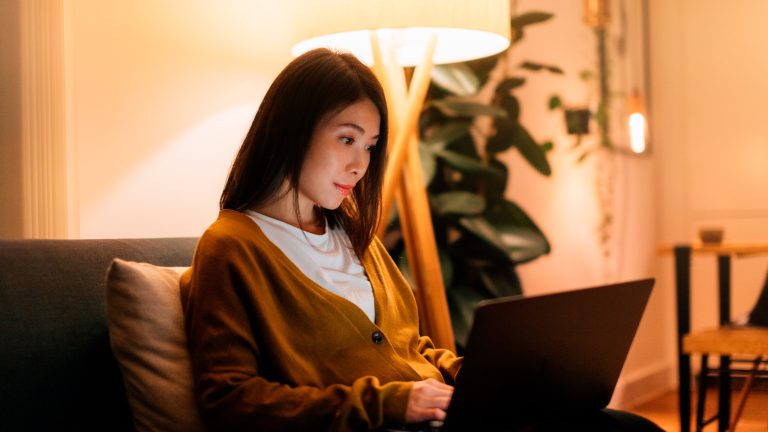 Eine Person sitzt bei künstlichem Licht auf einem Sofa vor dem Laptop.
