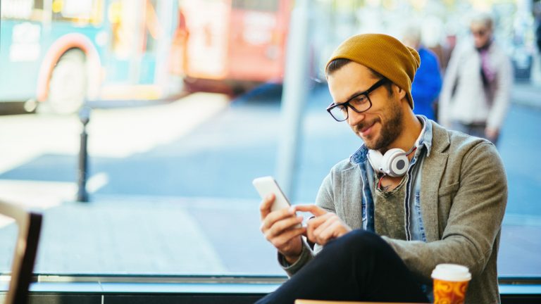 Eine Person sitzt in einem Café, trägt Kopfhörer um den Hals und schaut auf ihr Handy.