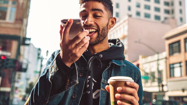 Eine Person läuft mit einem Kaffee in der Hand durch eine Stadt. Sie spricht dabei in ihr Smartphone.