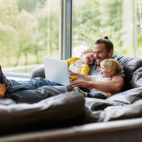 Ein Mann liegt mit seinen zwei jungen Kindern auf einer großen braunen Couch und sieht auf einem Laptop Videos.