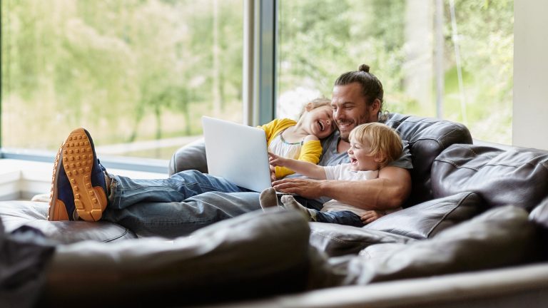 Ein Mann liegt mit seinen zwei jungen Kindern auf einer großen braunen Couch und sieht auf einem Laptop Videos.