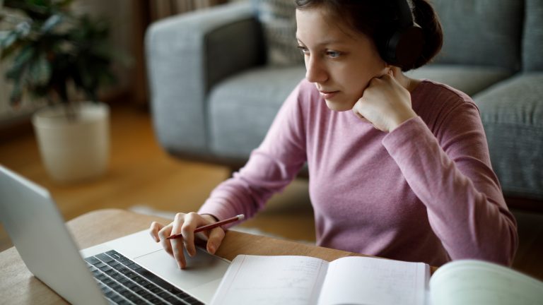 Eine Person sitzt mit einem Stift in der Hand vor einem MacBook. Daneben liegen aufgeschlagene Hefte. Eine Person sitzt mit einem Stift in der Hand vor einem MacBook. Daneben liegen aufgeschlagene Hefte.