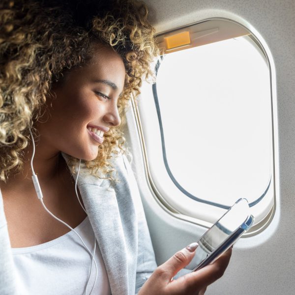 Eine Person mit voluminösen, lockigen Haaren sitzt am Fenster im Flugzeug und hört mit Kopfhörern Musik von ihrem Smartphone.