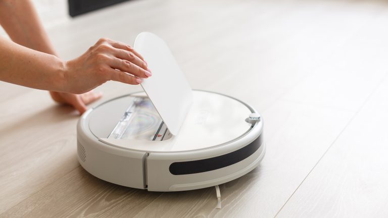 Eine Hand öffnet die Klappe an der Oberseite eines weißen, flachen, runden Wischroboters.