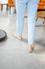 Ein schwarzer, kleiner, runder, flacher Wischroboter steht neben den Füßen einer Person in heller Jeans. Im Hintergrund stehen eine orange Couch und eine Couchtisch-Kombination aus Teakholz.