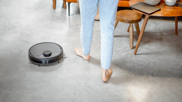 Ein schwarzer, kleiner, runder, flacher Wischroboter steht neben den Füßen einer Person in heller Jeans. Im Hintergrund stehen eine orange Couch und eine Couchtisch-Kombination aus Teakholz.