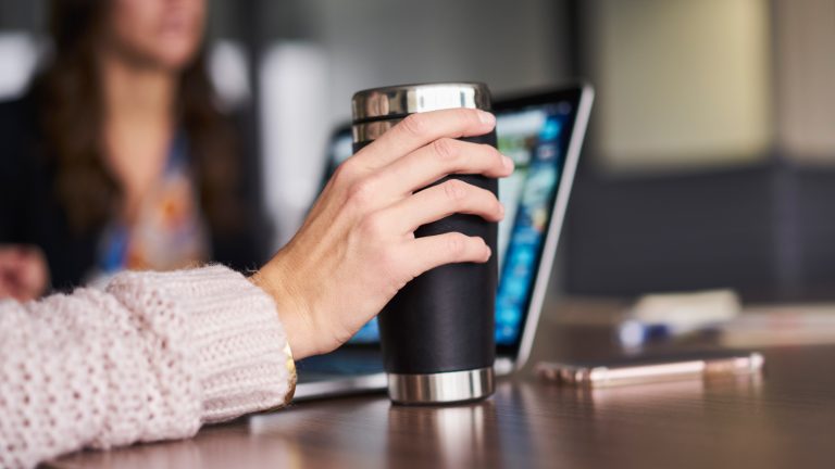 Eine Person sitzt an einem REchner in einem Büro und hält eine Thermoskanne in der Hand.