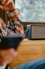 Eine Person mit Smartphone in der Hand lehnt an einer Holzkommode, auf der ein Bluetooth-Lautsprecher steht.