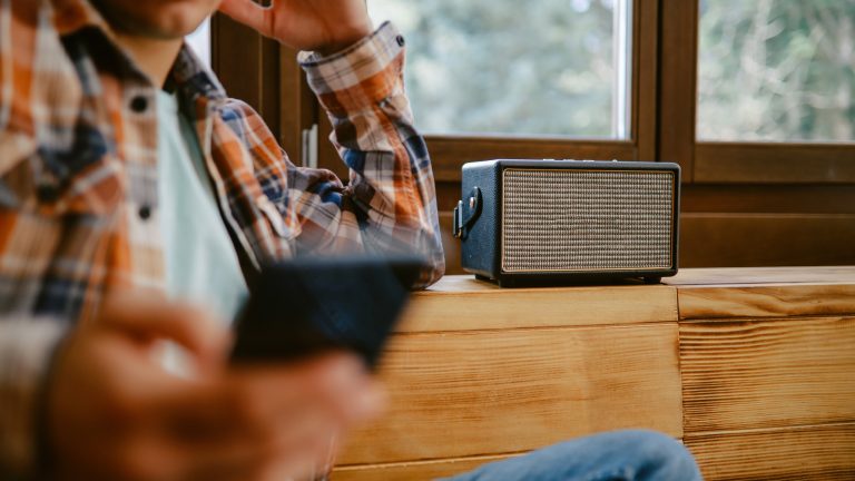 Eine Person mit Smartphone in der Hand lehnt an einer Holzkommode, auf der ein Bluetooth-Lautsprecher steht.