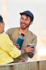 Zwei Personen stehen im Freien und lachen. Eine Person reicht der anderen eine Thermoskanne.