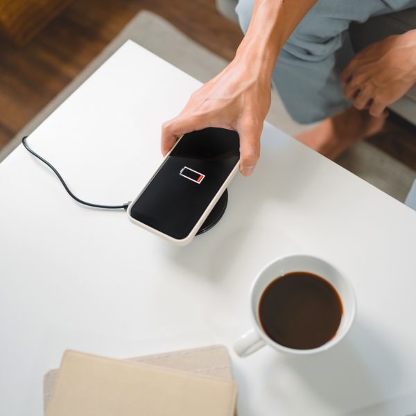 Eine Person in hellen Jeans legt ein Smartphone auf ein rundes wireless charging tool auf einem weißen Tisch. Daneben steht eine Tasse Kaffee.