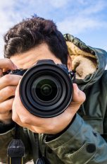 Eine Person mit kurzen braunen Haaren in einer dunkelgrünen Outdoorjacke hält eine Kamera vor sein Gesicht und fotografiert frontal.