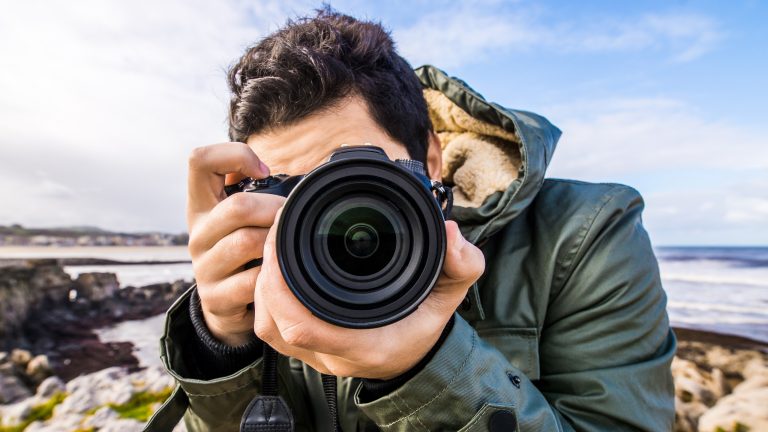 Eine Person mit kurzen braunen Haaren in einer dunkelgrünen Outdoorjacke hält eine Kamera vor sein Gesicht und fotografiert frontal.