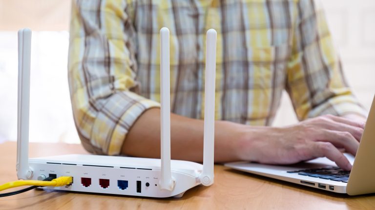 Ein Mann sitzt mit einem Laptop neben dem Router am Tisch.