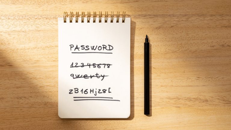 Notizblock auf einem Holztisch mit dem Begriff ‘PASSWORD’ oben und einer Liste von einfachen, durchgestrichenen Passwörtern wie ‘1234567’. Das letzte Passwort ist komplex und doppelt unterstrichen. Neben dem Notizbuch liegt ein schwarzer Stift.