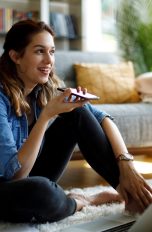 Eine Person sitzt auf einem Teppich vor einem Sofa. In der Hand hält sie ein Smartphone flach vor den Mund. Vor ihr steht ein aufgeklappter Laptop.