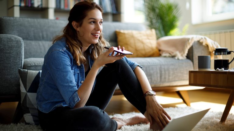 Eine Person sitzt auf einem Teppich vor einem Sofa. In der Hand hält sie ein Smartphone flach vor den Mund. Vor ihr steht ein aufgeklappter Laptop.