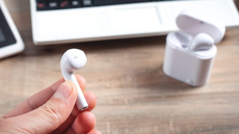 Eine Person hält einen AirPod in die Kamera. Im Hintergrund ist das dazugehörige Ladecase zu sehen.