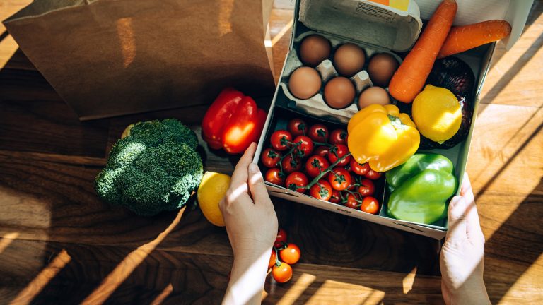 Hände präsentieren eine Auswahl an buntem Gemüse in einer weißen Pappbox auf einem hellen Holztisch.