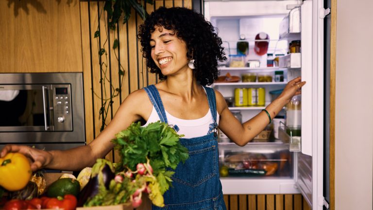 Eine Person mit schwarzen lockigen Haaren in einer dunklen Jeans-Latzhose räumt eine gelbe Paprika in einen geöffneten Kühlschrank.