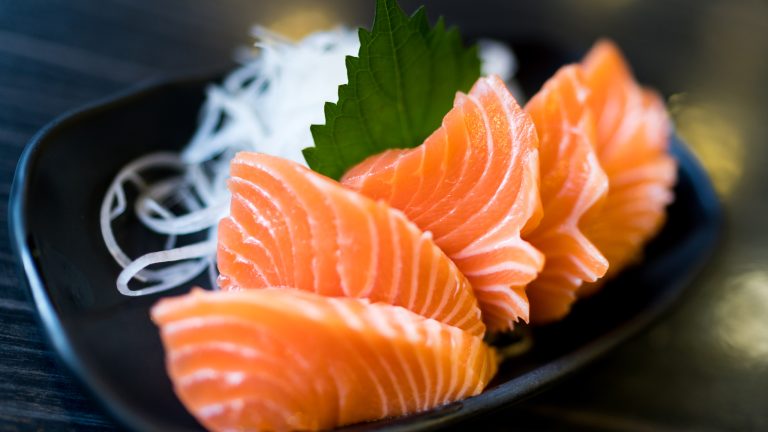 Frische Lachsstücke sind dekorativ mit Glasnudeln auf einem ovalen schwarzen Teller angerichtet.