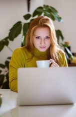 Eine Person mit einer Kaffeetasse in der Hand sitzt vor einem Laptop.