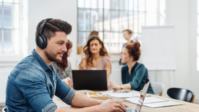 Eine Person sitzt mit Over Ear Kopfhörern an ihrem Laptop im Büro. Im Hintergrund sind Kolleg*innen zu sehen, die sich unterhalten.