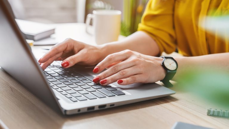 Eine Person tippt mit beiden Händen auf ihrer Laptoptastatur. Sie trägt eine gelbe Bluse, eine schwarze Armbanduhr und roten Nagellack. Eine Person tippt mit beiden Händen auf ihrer Laptoptastatur. Sie trägt eine gelbe Bluse, eine schwarze Armbanduhr und roten Nagellack.