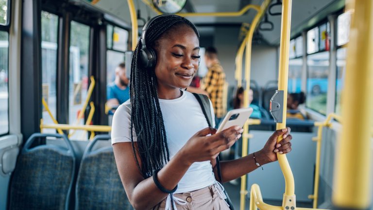 Eine Person mit vielen langen schwarzen Zöpfen steht im Bus und hört mit Over Ear Kopfhörern Musik von ihrem Handy.