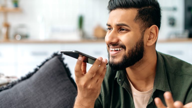 Ein Mensch sitzt in einem dunkelgrünen Hemd auf einer grauen Couch und telefoniert mit seinem iPhone in der Hand vor dem Mund.