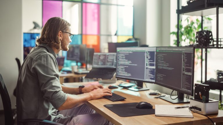 Eine Person sitzt an einem Schreibtisch, auf dem ein MacBook mit zwei angeschlossenen Monitoren zu sehen ist.