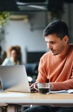 Ein Mensch in orangenem Pullover sitzt in einem Büro vor einem Laptop. Im Hintergrund sieht man andere Mitarbeitende und Pflanzen.