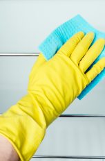 Eine Hand in einem gelben Gummihandschuh wischt die Ablageflächen in einem Kühlschrank mit einem blauen Schwammtuch. 