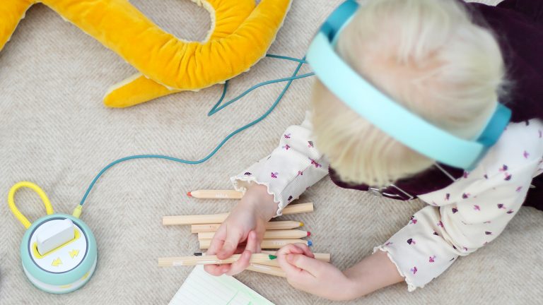 Galakto: Kinder-Lautsprecher ohne Internet, dafür mit Twist’n’Go Ein Kind mit Kopfhörern auf den Ohren liegt auf dem Fußboden und ist mit Buntstiften beschäftigt, die vor ihm liegen. Daneben liegt der Player des Galakto, an dem der Kopfhörer angeschlossen ist.