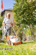 Ein Mensch mit weißem T-Shirt mäht mit einem weiß-orangenen Akkurasenmäher seinen Garten. Die Sonne scheint und der Rasen und die Pflanzen sind grün.
