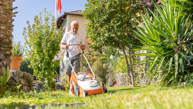 Ein Mensch mit weißem T-Shirt mäht mit einem weiß-orangenen Akkurasenmäher seinen Garten. Die Sonne scheint und der Rasen und die Pflanzen sind grün.