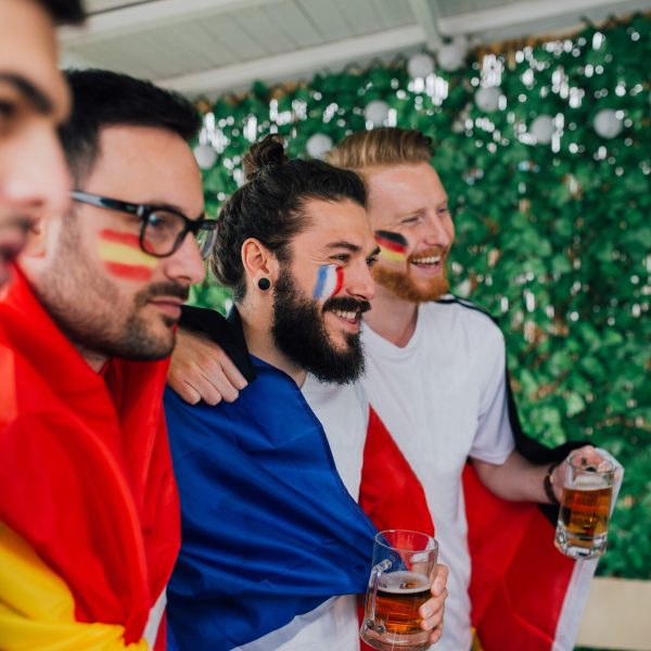Vier Personen mit Länderfahnen schauen in Richtung eines nicht sichtbaren Fernsehers.