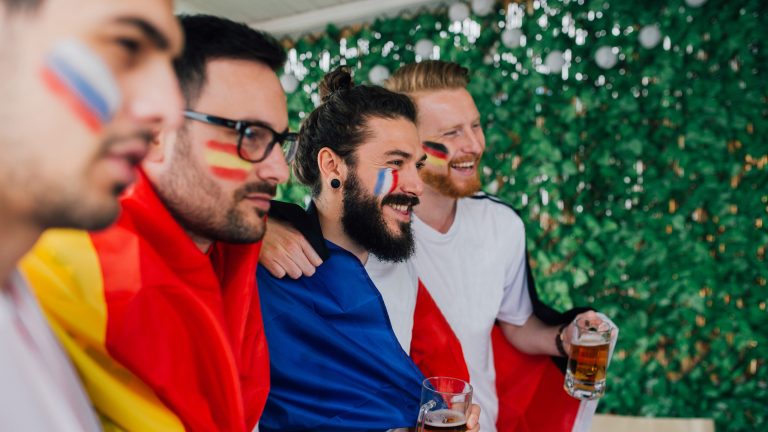 Vier Personen mit Länderfahnen schauen in Richtung eines nicht sichtbaren Fernsehers.