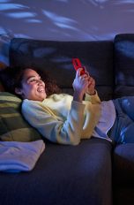 Eine Person liegt im dunklen Wohnzimmer auf der Couch und scrollt an ihrem Handy.