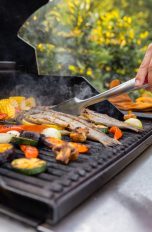 Eine Person steht mit einer Grillzange an einem Gasgrill und wendet Fisch.