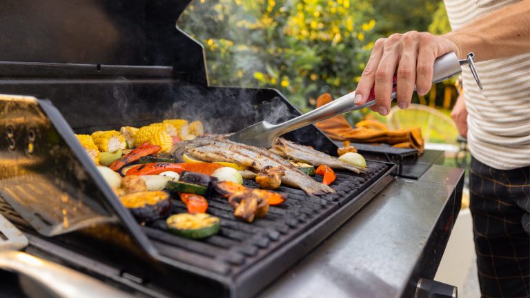Eine Person steht mit einer Grillzange an einem Gasgrill und wendet Fisch.