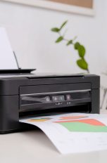 Ein Drucker steht neben einem Laptop und einer Pflanze auf einem Tisch und druckt ein Blatt mit bunten Mustern.