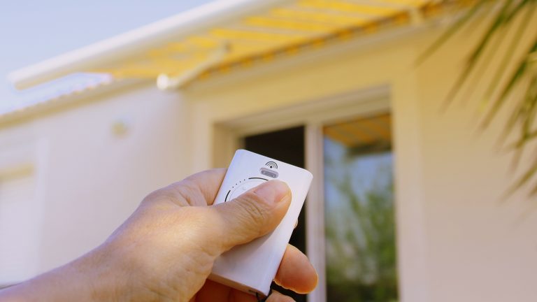 Markisenmotor nachrüsten Eine Person hält die Funkfernbedienung einer Markise in der Hand. Im Hintergrund ist die Markise über einem Terrassenfenster zu sehen.