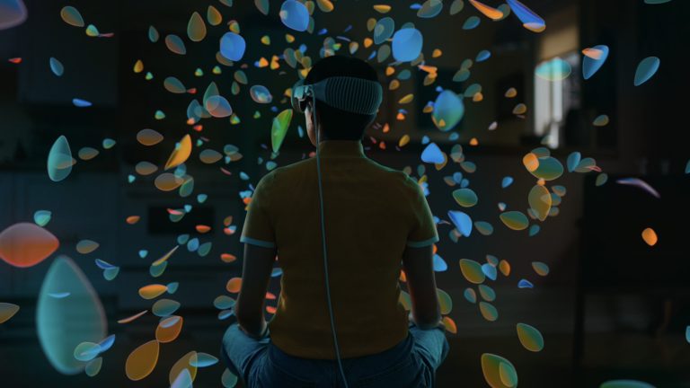 Eine Person sitzt in ihrer Wohnung auf dem Boden und trägt eine Apple Vision Pro. Um sie herum schweben Blütenblätter in einer 3D-Welt Eine Person sitzt in ihrer Wohnung auf dem Boden und trägt eine Apple Vision Pro. Um sie herum schweben Blütenblätter in einer 3D-Welt.