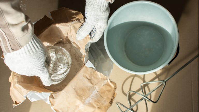 Fliesenfugen ausbessern Eine Person nimmt etwas Pulver für die Fugenmasse aus einer Verpackung. Neben ihr steht ein Eimer.