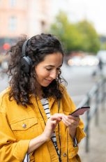 Eine Person ist im Freien unterwegs, trägt Kopfhörer und bedient dabei ihr Smartphone.