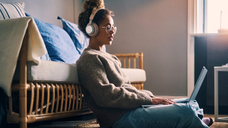 Spotify, So nutzt du es unter iOS und Android Eine Person sitzt vor einem Sofa auf dem Boden, trägt Kopfhörer und hat einen Laptop auf dem Schoß.