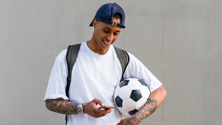 Eine Person mit einem Fußball unter dem Arm schaut auf ihr Smartphone.