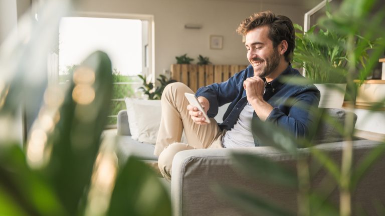 TikTok Coins, so lädst du sie auf und verteilst Geschenke Eine Person sitzt in einem Wohnzimmer mit vielen Pflanzen auf der Couch und scrollt auf ihrem Smartphone.